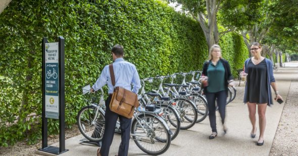 A business man with a leather messenger bag renting a bike from Zagster bike share while two women with drinks walk past