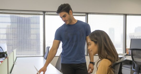 A man and woman in an open space office looking over a book with an open laptop, glass of water, and smartphone on the desk