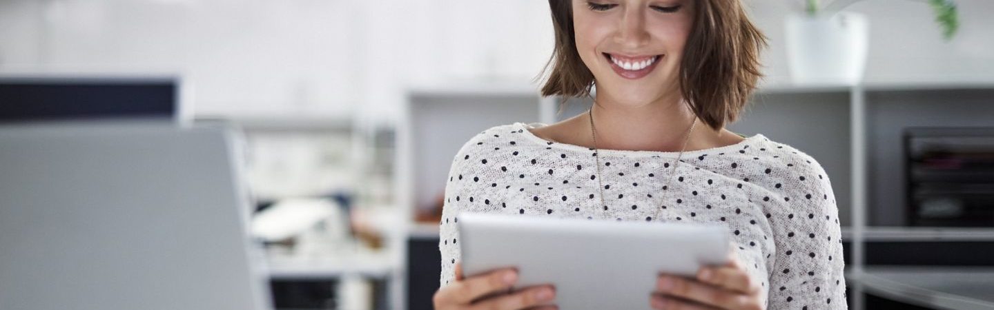 A smiling young woman in a spotted blouse looking at a tablet with a laptop and other office supplies on the desk in front of her