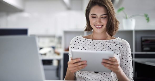 A smiling young woman in a spotted blouse looking at a tablet with a laptop and other office supplies on the desk in front of her