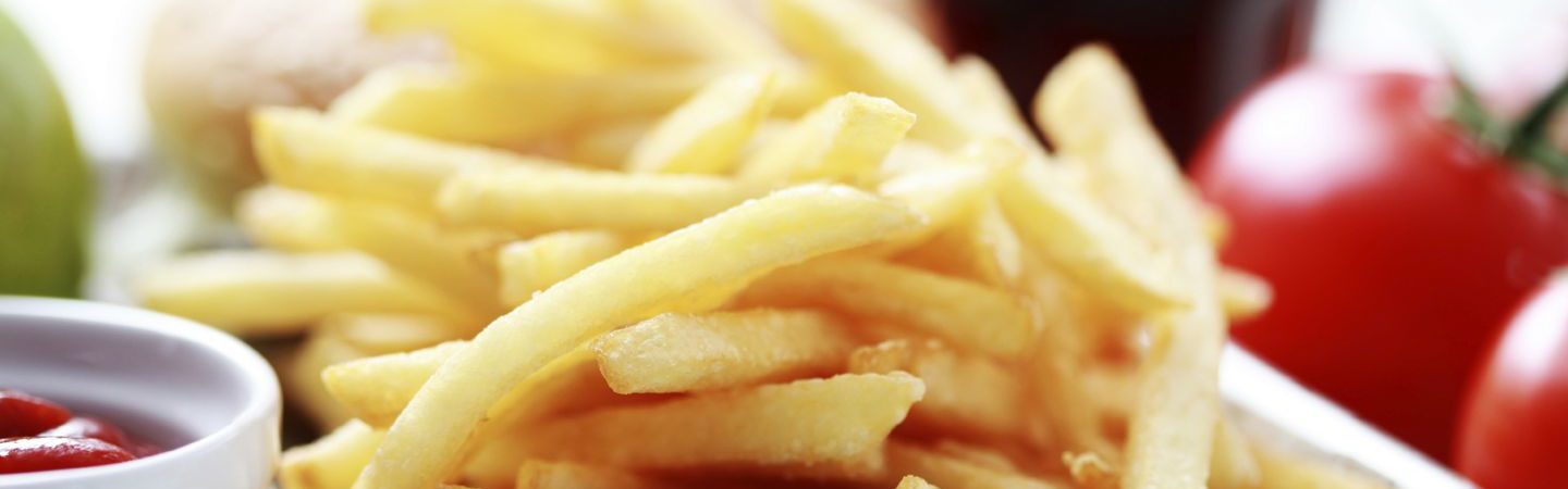 French fries in a square bowl and small dipping bowl containing ketchup with tomatoes in the background.