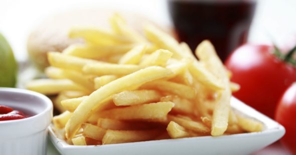 French fries in a square bowl and small dipping bowl containing ketchup with tomatoes in the background.