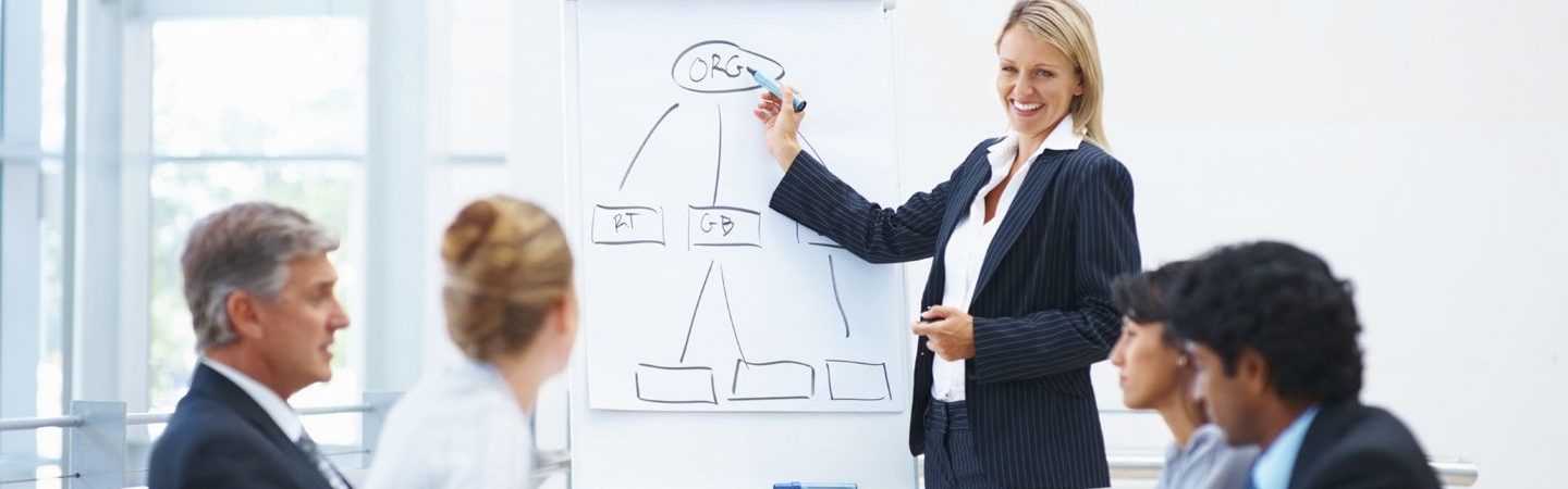 A group of businesspeople sitting together in a meeting while a woman is standing and drawing a diagram in marker on a large notepad