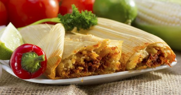 Tamales on a plate with a red pepper, lime wedge, cilantro and tomatoes, corn, and lime in the background.