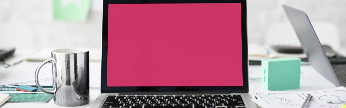 Laptop with a bright magenta screen sitting on a desk with a cutting board, coffee mug, and other office supplies