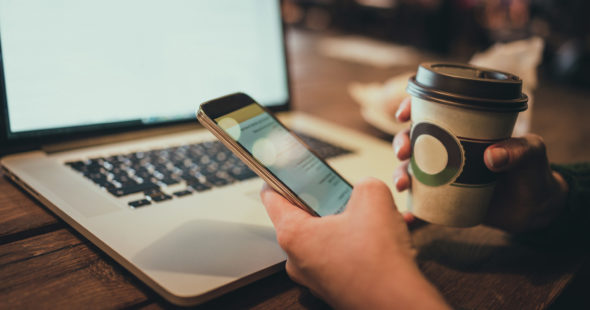 A person holding a cup of coffee and using their cellphone while sitting in front of their laptop